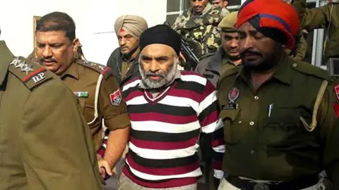 Bharat Bhushan/Hindustan Times via Getty Images Former KLF leader Harminder Singh Mintoo being brought to court in Patiala, India, for interrogation in December 2016. He is surrounded by men in uniform and is weaging a white, black and maroon striped jumper.