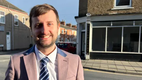 A man with short brown hair and a short beard, and wearing a grey suit and blue striped tie, smiles at the camera as he stands in Pasture Road, Goole, in front of a shop.