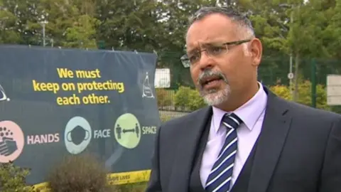 Craig D'Cunha, who has greying hair, a beard and glasses, and is wearing a jacket and tie, standing in the school grounds