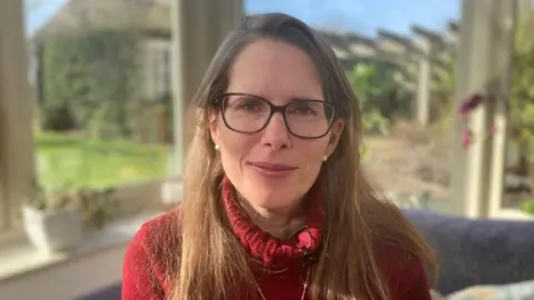 Georgie Maynard is wearing a red jumper and sitting in her conservatory at home