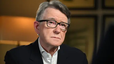 BBC Lord Mandelson in front of a dark orange background looking serious