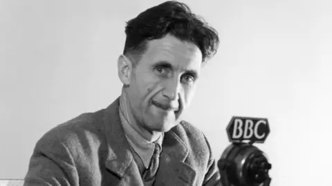 A black and white photo of George Orwell who is smiling at the camera in front of a microphone that has the BBC logo on top of it. He has dark hair and wears a suit jacket, shirt and tie.