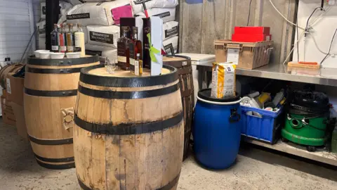 Three wooden whisky barrels with glass bottles on them, which are full of different liquor like whisky and vodka. Behind there are white bags of barley piled up. On the right is a metal preparation table with various equipment around including a vacuum. 