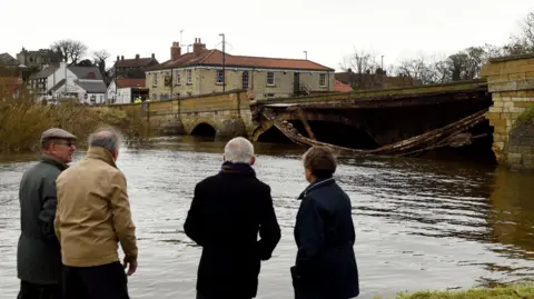 PA Media Residents view damage to Tadcaster's bridge following heavy flooding over Christmas in 2015. A section of the bridge has collapsed into the river, with contractors working at the far end of the bridge.