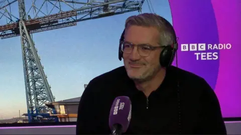 Craig McManus from the MFC Foundation is sitting in the purple-themed Radio Tees studio. He is wearing headphones and speaking into a microphone.