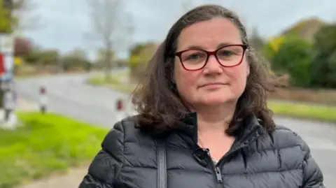 Keena Entwistle pictured outside with a road in the background. She has dark/greying hair, red rimmed glasses and is wearing a black puffer jacket. 