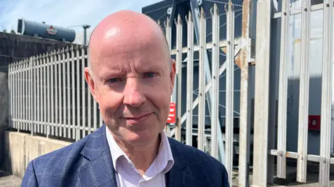 A man looking into the camera. He has a bald head and is wearing a blue and white checked blazer over a white shirt undone at the top button. He's outside on a sunny day and behind him is some metal fencing. Behind that, you can catch a glimpse of more grey metal, which is a substation.