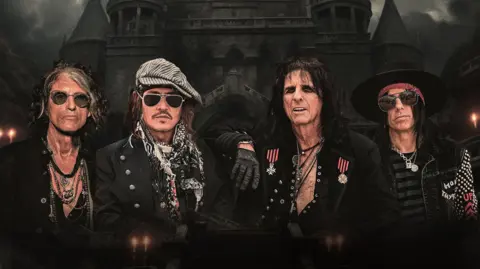 A group of four men dressed in dramatic rock‑and‑roll attire stand in front of a dark, gothic castle with tall spires. They wear sunglasses, hats, scarves, jewellery, and studded jackets, with the central figure in a medal‑covered coat.