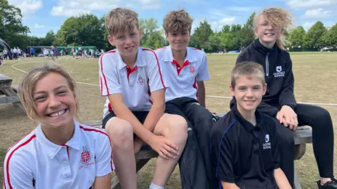 Three pupils from Kingswood School on the left in white cricket shirts, and two pupils from Oldfield School on the right, in black cricket shirts. There is a cricket pitch behind them.