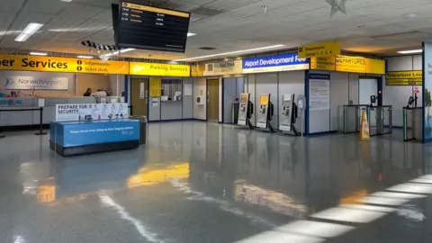 Shaun Whitmore/BBC The inside of an airport terminal, with yellow illuminated signs.