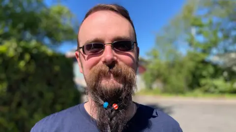 A head and shoulders photograph of William Hall who has a long beard with beads in it. He is wearing a navy tshirt.