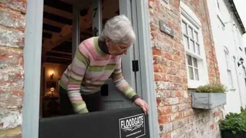 An older woman with grey hair and stripy jumper attaches a black rectangular barrier to her front door which says "Flood Gate". Her home is red-brick and terraced.