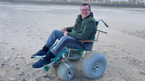 Shane Robins in a beach accessible wheelchair on Rhyl beach 