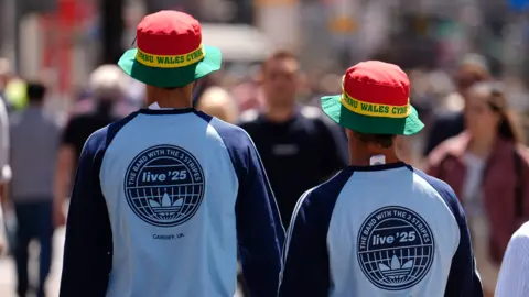 Two men in Oasis shirts which say 'the band with three stripes' on the back. The shirts are light blue with blue sleeves. They're also wearing Wales bucket hats which are red yellow and green. They're walking towards a crowd of people