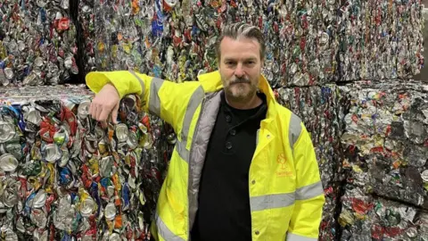 A man in a high-vis jacket leaning on, and standing in front of, bales of metallic recycling - mainly crushed cans.