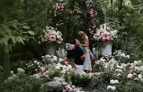 @TAYLORSWIFT Taylor Swift y Travis Kelce en un fondo floral.
