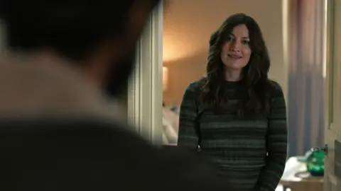 Netflix A woman in a patterned sweater stares at a man in a domestic setting who is standing with his back to the camera