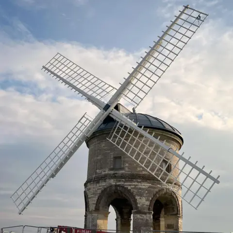 Four white sails in position on a round windmill, with arches at the foot of it.