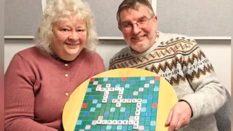 Helen is wearing a peach pink cardigan with golden buttons holding a scrabble board that has words Helen, Twenty Years, Graham, Married, Scrabble fitted in the scrabble board and Graham holding the Scrabble board as well and is wearing a crème coloured winter jumper. 