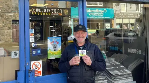 David Thomas Photo of a man with a silver beard and a fred perry black baseball cap on. He stands outside a carpet store in the sunlight, holding a coffee and a "no to the gate" sticker. In the window behind him a "No to the gate" poster can be seen. 