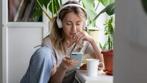 Getty Images A stock shot of a young blonde woman wearing white headphone and looking at her phone.
