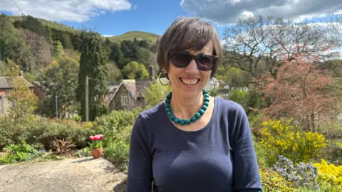 A woman wearing a blue top and large green necklace is smiling at the camera. She is standing in a sunny garden with large hills in the distance. She is wearing sunglasses and hooped earrings.