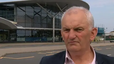 Aviation group member Joe Mooney - A middle aged white man with grey hair wearing a pink shirt and navy jacket stood outside the airport