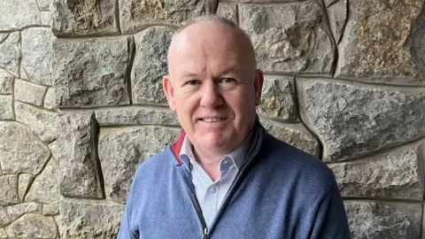 Aled Morris Jones stood in front of a wall. It is a head and shoulders shot. He is balding and has a blue zip up sweater on, over a light blue shirt.