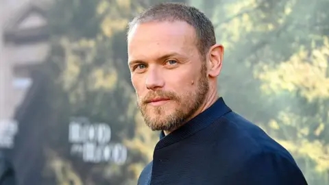 Sam Heughan looking slightly off camera. He is wearing a blue jacket over a dark blue top and is standing in front of a blue background with yellow writing on it.