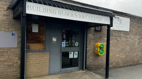 The entrance to a community hub, it has a sign saying Building Blocks Day Centre CIC. There is a defibrillator in a yellow and green case on the wall outside.