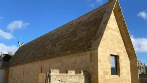 The Faith Museum in Bishop Auckland is a light stone building. The image is taken from the side and shows the side and one end of the building with a window in the end wall. The image also shows some of the building behind and a bright blue sky with some patchy clouds