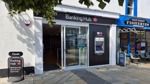Cockermouth Banking Hub. The facility has a glass entrance door and a cashpoint. A sign for the hub is also on the pavement.