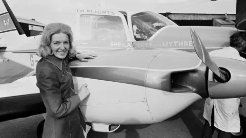 Express / Getty The woman from the above images, again in black and white, standing next to a twin-engined light aircraft with "Sheila Scott and Mythre" emblazoned on the side. She is resting her hand on one of the engines.