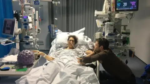 Bryony Thomas lies in a hospital bed hooked up to numerous machines as she battles Pancreatic Cancer, her husband leans on the side of the bed