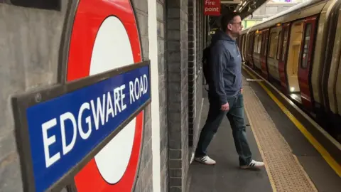 Ben Thwaites walking towards a train on the London Underground platform. The train doors are open. A blue and red sign in the foreground reads Edgware Road.