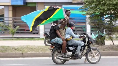 Three men on a motorbike, one is standing up and holding a big Tanzanian flag.