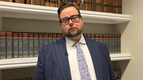A photograph of a man in a suit with classes and a beard in front of a book shelf showing a a large amount of old books.