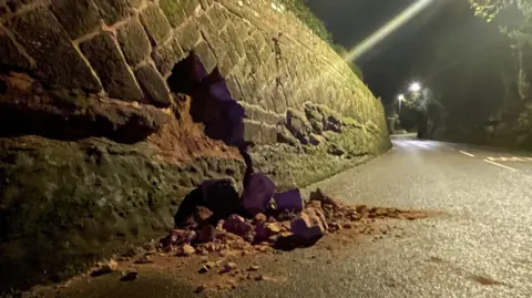 Stones in the road near a failed wall flush to the roadway