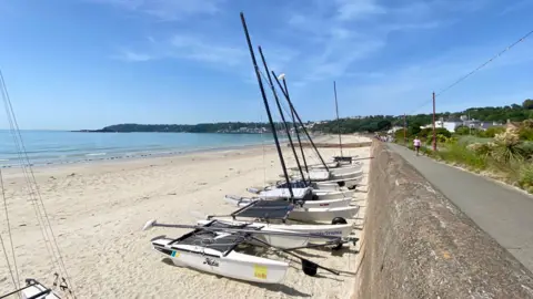 A row of small sailing boats sits on a sandy beach beside a coastal path, with calm blue sea, a distant headland and scattered houses under a bright, lightly clouded sky.