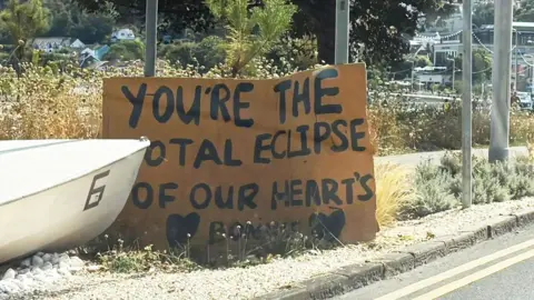 BBC A cardboard sign welcoming visitors to Mumbles reads "You're the Total Eclipse of our hearts Bonnie", referencing the singer's 1982 hit.