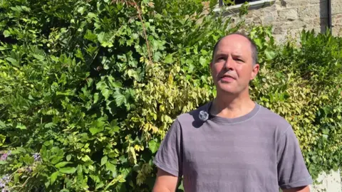 David Jones wears a purple T-shirt and stands in front of a hedge