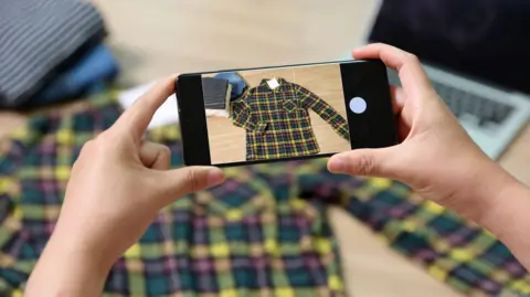 Getty Images A person's hands holding a phone showing a photo of a checked shirt, which you can see is under the phone lay on a desk, looking less in focus