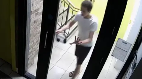 Coroner Service A screenshot of a youtube clip showing Noah Donohoe wearing black and white striped flip flops, dark shorts and a white t-shirt. He is holding a pair of black over the ear headphones and is pushing a door open. 