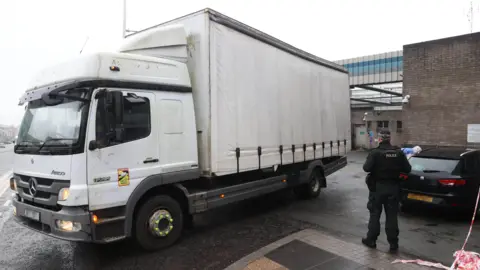 PA Media A large white lorry reverses into Lurgan police station. A police officer stands with his back to the camera and a forensic officer leans onto a black car.