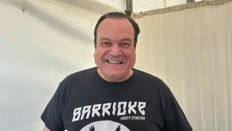 Shaun Williamson, a middle-aged man with dark hair. He is wearing a black t-shirt with the word 'BARRIOKE, PARTY STARTER' on it. 