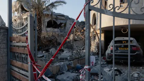The bomb site in Tamra, where rubble lies strewn across the driveway of a home, the gates held shut by red incident tape