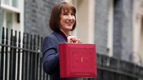 Reuters Chancellor Rachel Reeves standing outside Downing Street holding up a red briefcase that says "Chancellor of the Exchequer" and the Royal cypher. In gold writing. Rachel Reeves is smiling, with short brown hair and wearing a navy suit. 