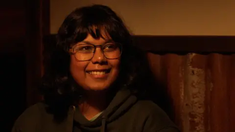 Swati Padmanabhan smiles just to the right of the camera. She is wearing a dark green hoodie and sitting in front of a brown and yellow wall. 