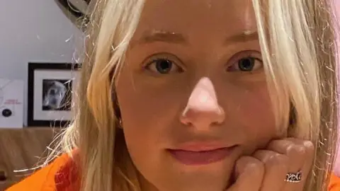 A girl called Kezia Mason. She has long, straight blonde hair. Her face is leaning against her left hand, which has a silver ring on her middle finger. She has a small smile on her face.