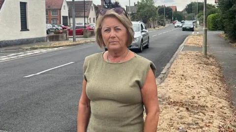 Elaine Kent a woman stood in a green top with green and white trousers beside a road with cars on it.